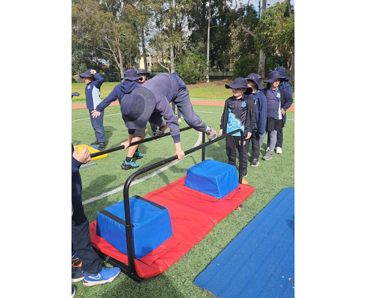 Students are outside on a sports field, actively participating in a fitness activity that includes running and jumping over obstacles, promoting fun and physical fitness.