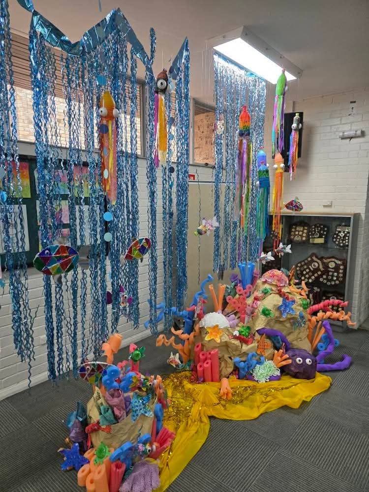 An art show display features a vibrant collection of student-created artworks, including sculptures and mixed media pieces, arranged creatively with blue hanging decorations and colorful figures, celebrating imagination and artistic talent within the school community.