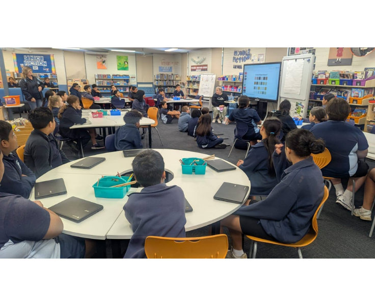 A group of students are seated at tables in a classroom, using tablets and listening to a teacher, demonstrating the use of technology and digital learning tools.