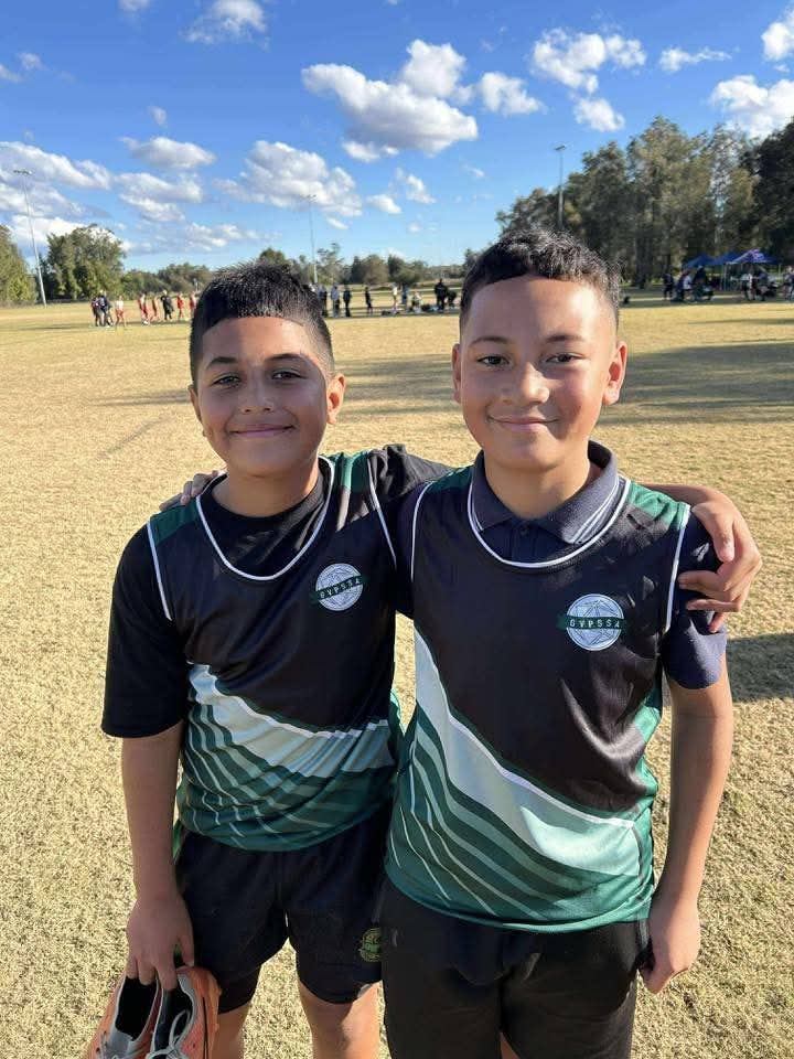 Two students stand side by side on a grassy sports field, wearing matching sports uniforms, representing their participation in Athletics Carnivals and GALA days at Cartwright Public School.