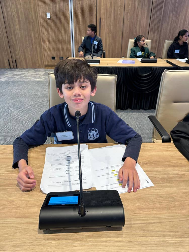 A student sits at a conference table with a microphone and notes, actively participating in the 2168 Student Parliament program, which empowers students to address school and community issues.